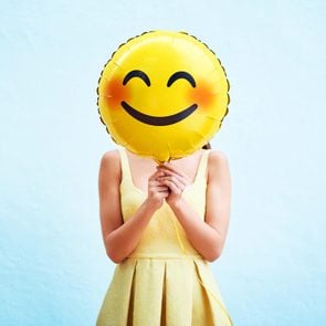 person in a yellow dress holding a blushing smiling emoji balloon in front of her face on a blue background