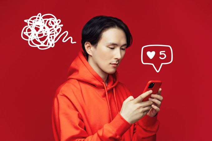 Serious Asian man wearing hoodie looking at and using smart phone on red background. Doodle of social media notification and scribbles