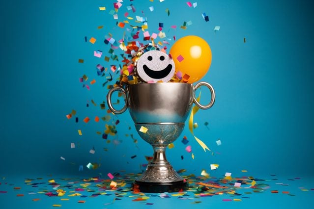 gold trophy on blue background with smiley face, baloon, and confetti coming out the top of the trophy.