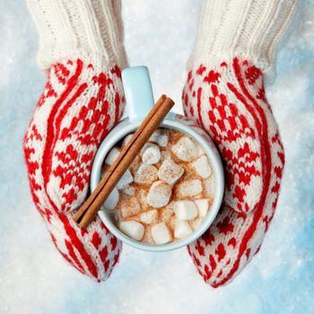 Hands wearing mittens holding a mug of hot chocolate with marshmallows and a cinnamon stick
