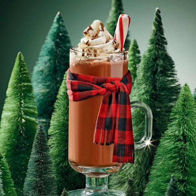 A glass of hot chocolate with a scarf and peppermint stick among plastic fir trees