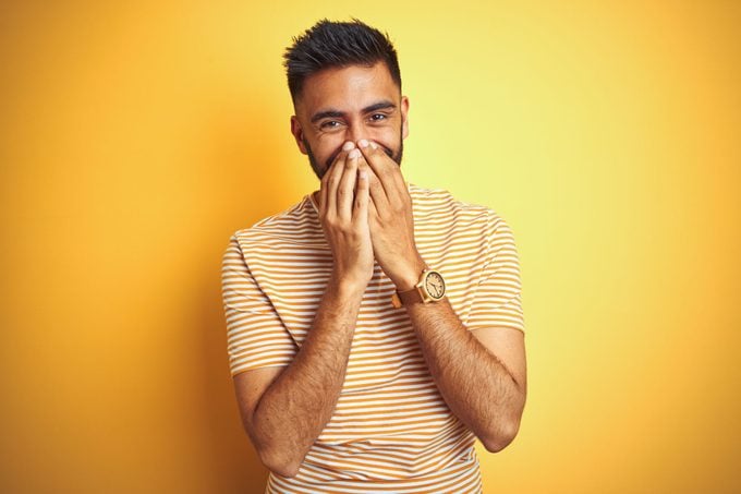 Young Indian Man Wearing a striped tshirt standing in front of a yellow background blushing and covering his face with his hands