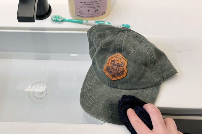 hand washing a baseball hat in a bathroom and dabbing with a microfiber cloth with a toothbrush and detergent in the background