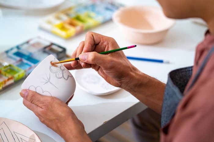 Asian LGBTQ guy painting self-made pottery at home studio.
