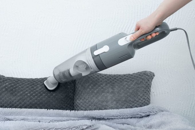 Handheld vacuum cleaner in hand. Vacuums fur decorative pillows on bed.