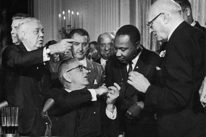 US President Lyndon B Johnson shakes the hand of Dr Martin Luther King Jr (1929 - 1968) at the signing of the Civil Rights Act while officials look on, Washington DC. (Photo by Hulton Archive/Getty Images)