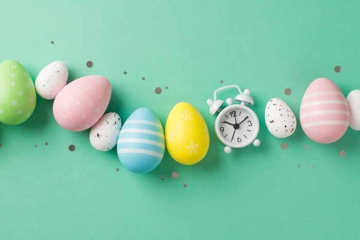 Top view photo of easter decorations glowing confetti row of multicolored easter eggs and white alarm clock on isolated turquoise background with copyspace