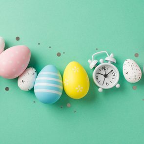 Top view photo of easter decorations glowing confetti row of multicolored easter eggs and white alarm clock on isolated turquoise background with copyspace