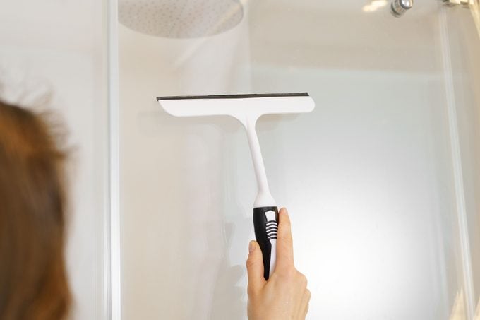 Close-up of a young woman using a scraper to remove drops from a glass shower and lime scale. The concept of cleaning in the apartment and in the bathroom