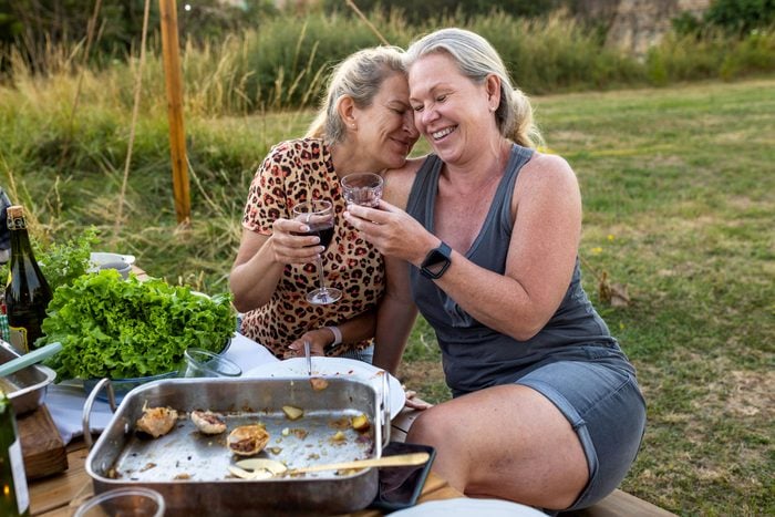 Two older woman are sitting at a dining table after a meal, they are holding wine glasses and are giggling with each other