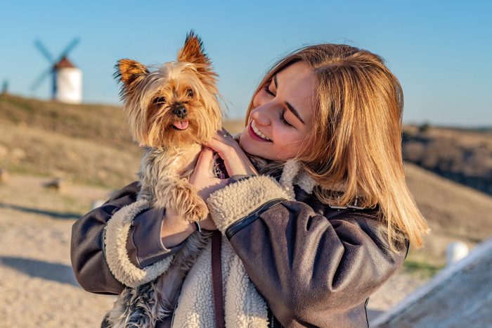 Young woman caressing a Yorkshire Terrier she's holding in her arms. Positioned in a rural setting on top of a hill surrounded by windmills.