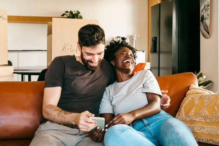 Happy multiracial couple enjoying sharing smart phone while sitting on sofa in living room at home