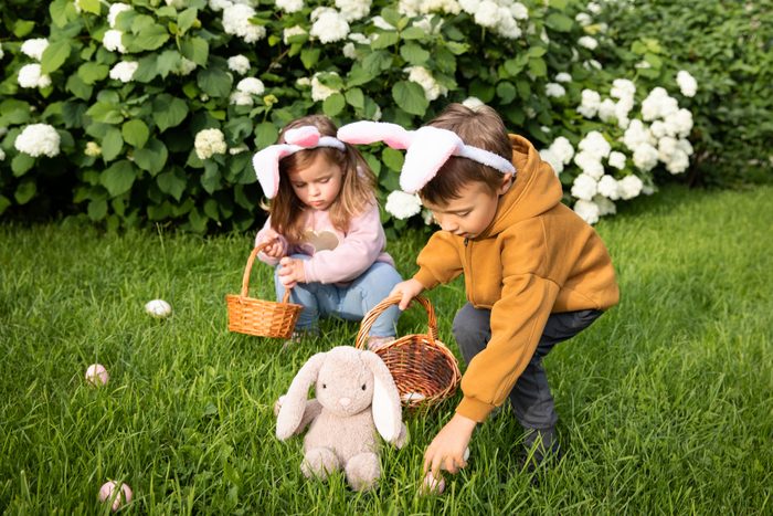 Little boy and girl in costumes with bunny ears hunt for eggs in the spring garden on Easter day.