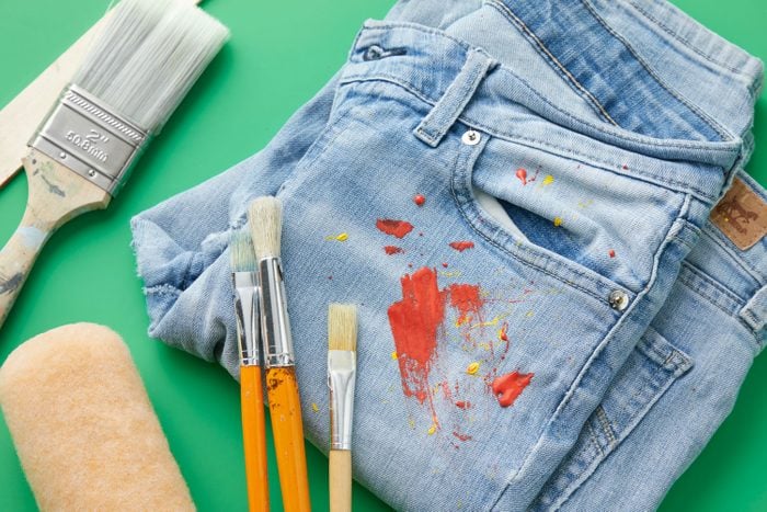 Paint stains on a pair of jeans with artist and household paintbrushes nearby on green background