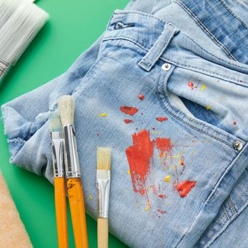 Paint stains on a pair of jeans with artist and household paintbrushes nearby on green background