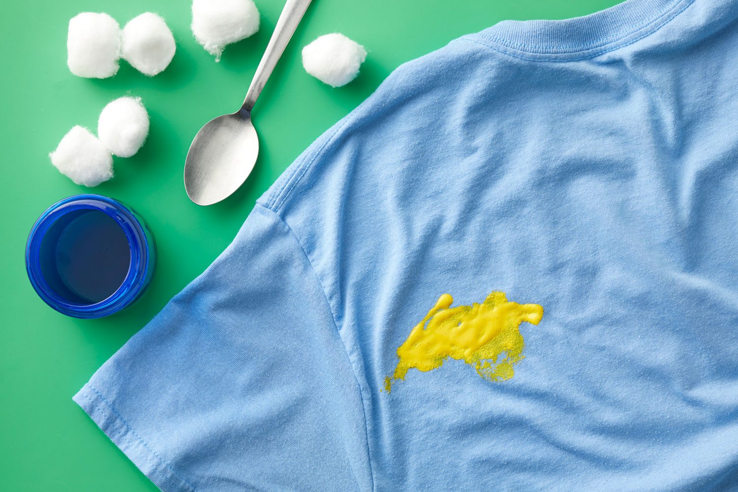 Yellow acrylic, water-based paint stain on a blue t-shirt on green background with supplies needed to clean it nearby