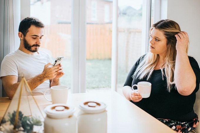 a couple sitting at a kitchen table. The man is on his phone and the woman is playing with her hair drinking a cup of coffee