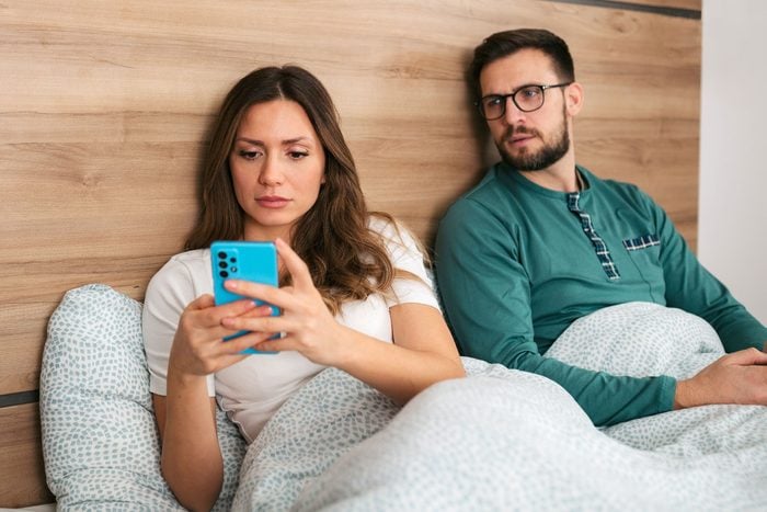 a woman is on her phone in bed and her suspicious boyfriend is looking over her shoulder wondering if she is cheating