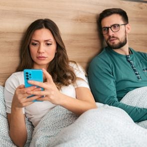 a woman is on her phone in bed and her suspicious boyfriend is looking over her shoulder wondering if she is cheating