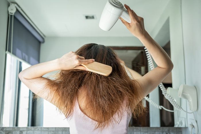 Rear view of young Asian woman using a comb for brushing her hair with a hair dryer for blowing water to dry her hair.