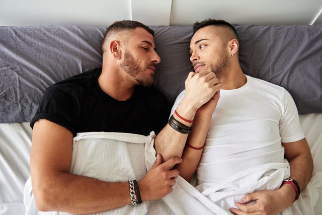 Man looking at his sleeping boyfriend while they holding hands in bed.